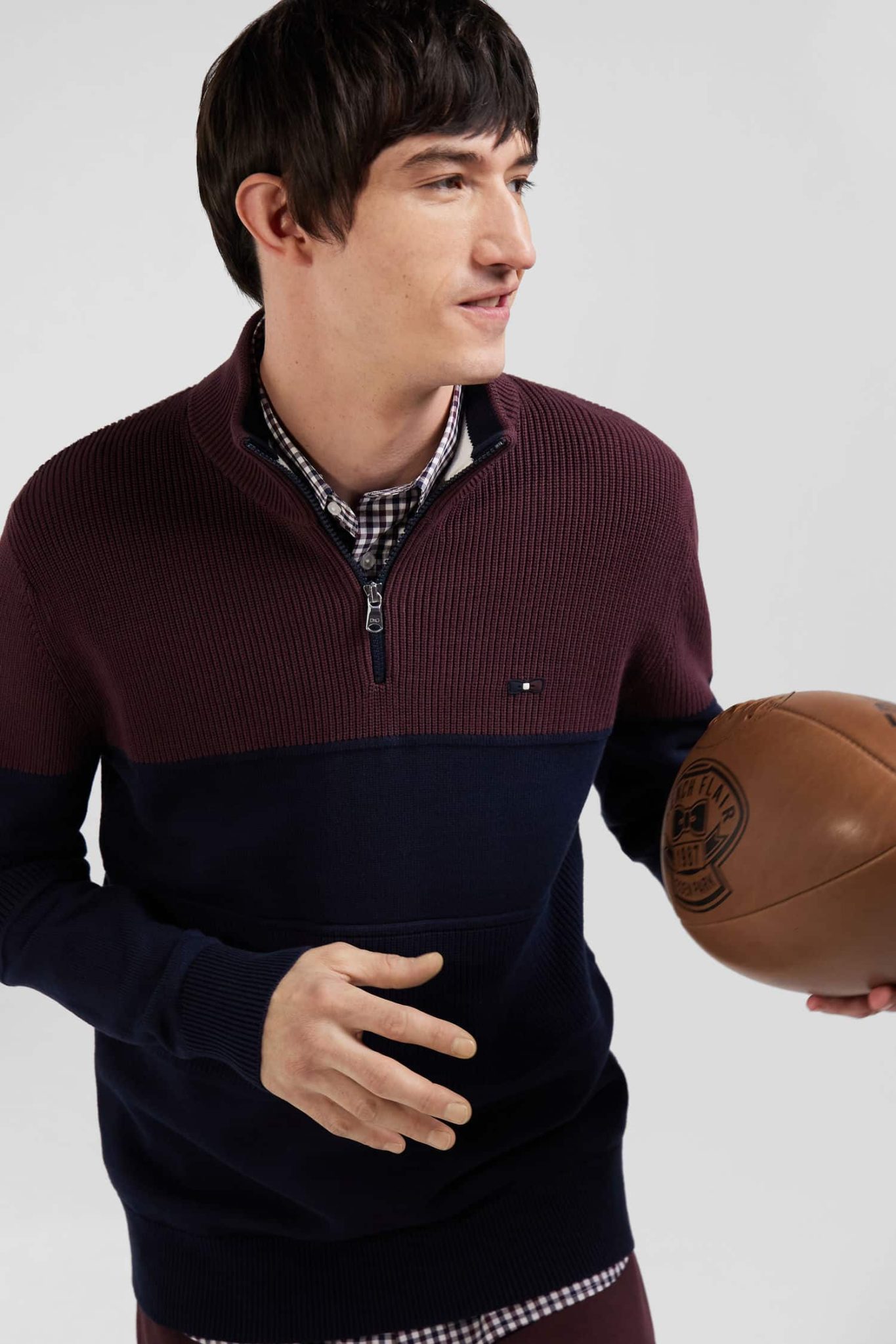 A person wearing a maroon and navy sweater with a half-zip is holding a brown basketball under one arm. They are looking to the side, standing against a plain background.