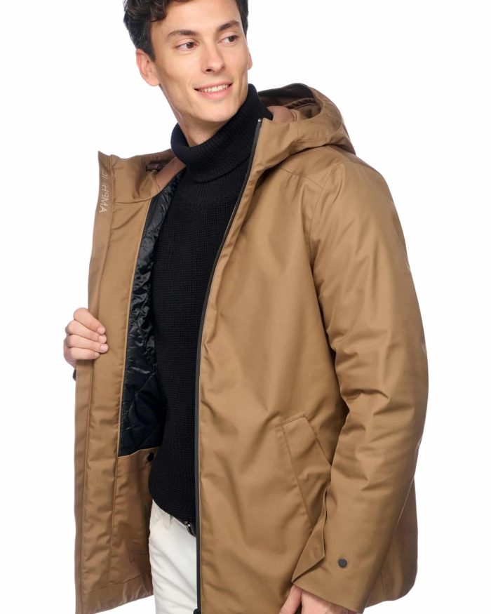 A person wearing a brown hooded jacket over a black turtleneck and light-colored pants stands against a white background. They are smiling and holding the open front of the jacket with one hand, revealing part of the black interior lining.