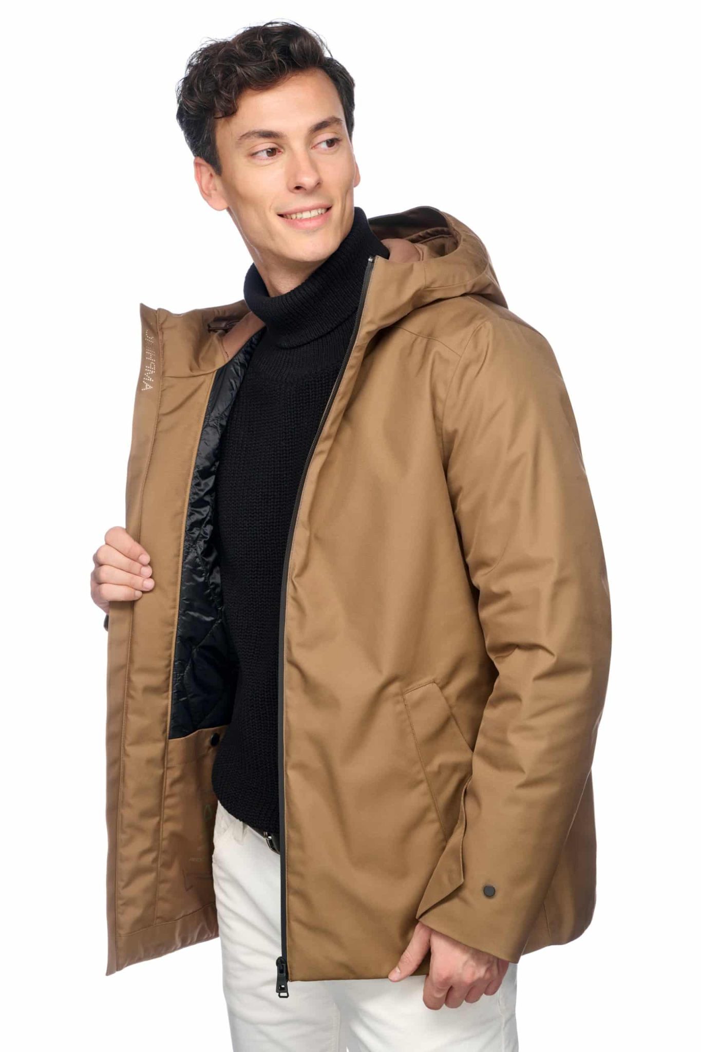 A person wearing a brown hooded jacket over a black turtleneck and light-colored pants stands against a white background. They are smiling and holding the open front of the jacket with one hand, revealing part of the black interior lining.