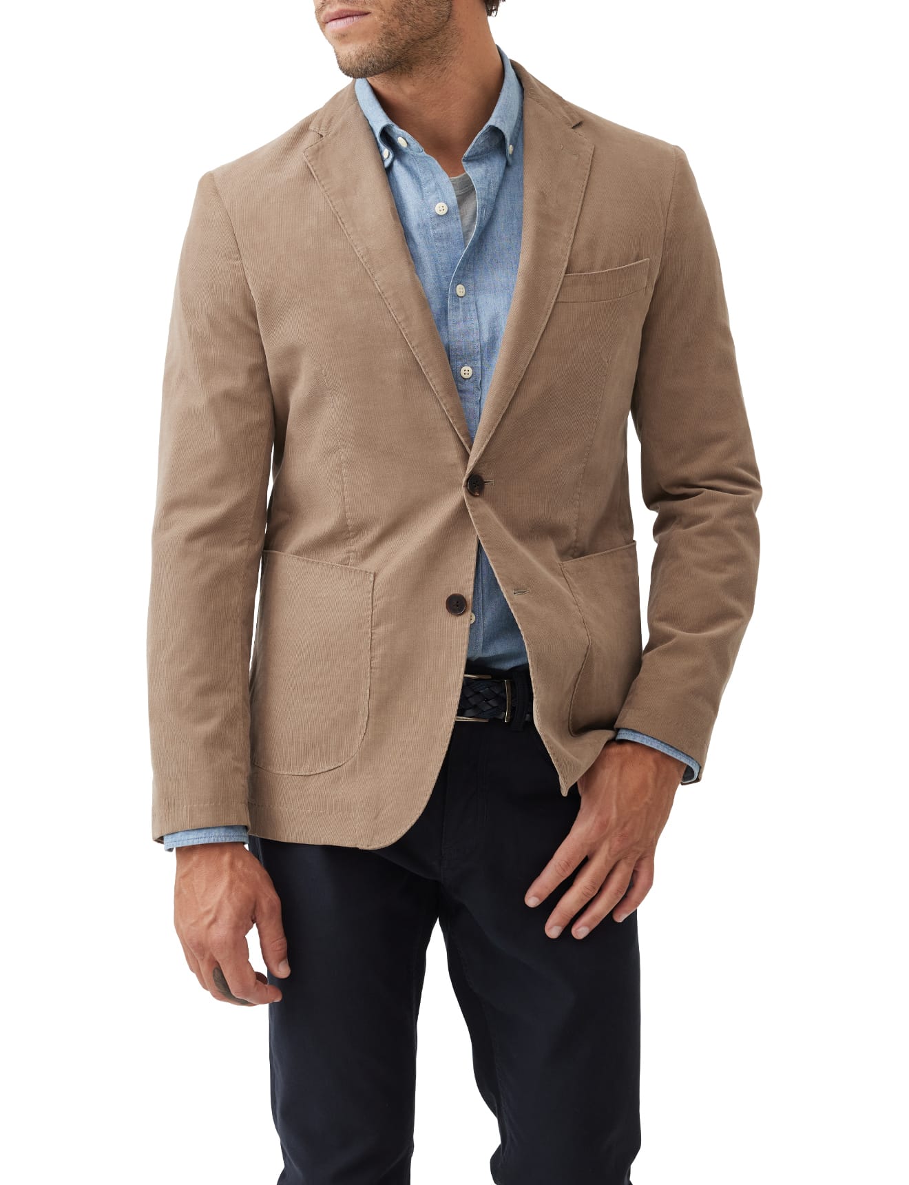 A man wearing a tan blazer over a light blue button-up shirt paired with dark pants stands against a white background. His left hand is resting casually by his side, and his right hand lightly touches his waist.