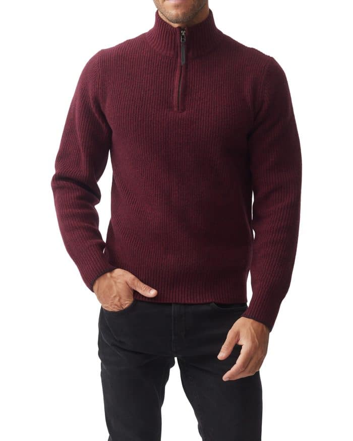 A person wearing a burgundy ribbed sweater with a half-zip collar stands against a white background. They are also wearing dark pants, with one hand in their pocket.