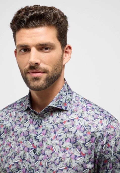 A man with brown hair and a beard is wearing a purple and green floral-patterned shirt. He is looking at the camera with a neutral expression. The background is plain and light-colored.