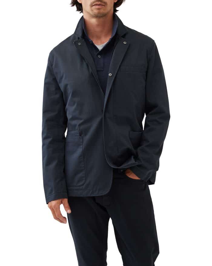 A person wearing a dark blue blazer over a navy shirt and black pants stands against a plain white background. The blazer has a buttoned collar and flap pockets, and the person has their right hand in their pocket.