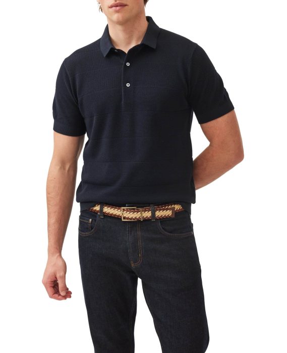 A person wearing a black short-sleeve polo shirt, dark jeans, and a woven belt. The shirt has a textured pattern, and the person’s left hand is tucked behind their back. The background is plain white.