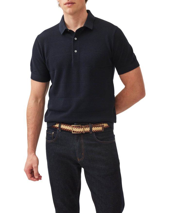 A person wearing a black short-sleeve polo shirt, dark jeans, and a woven belt. The shirt has a textured pattern, and the person’s left hand is tucked behind their back. The background is plain white.