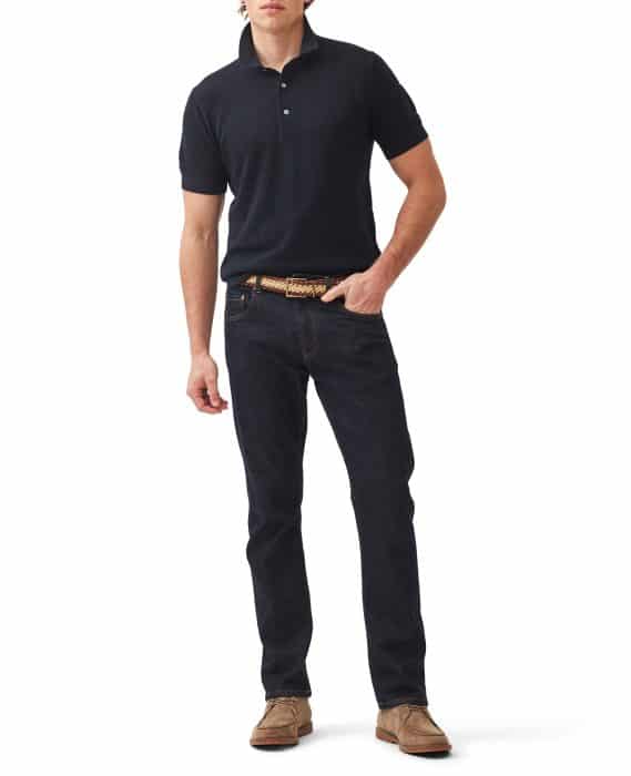 A person stands against a white background, wearing a black polo shirt, dark blue jeans, a woven beige belt, and brown suede shoes. One hand is in their pocket, and they have a relaxed posture.