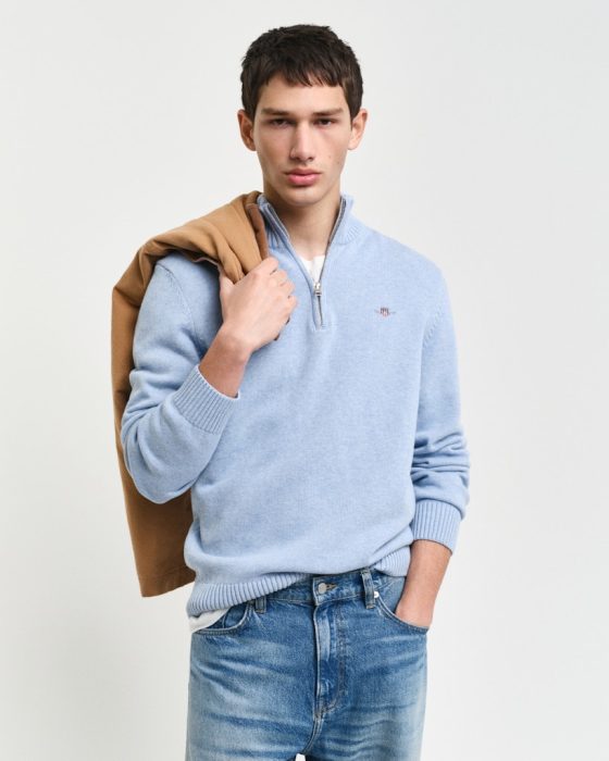 A person wearing a light blue sweater and jeans with one hand in their pocket. They have a brown jacket draped over their shoulder against a plain background.