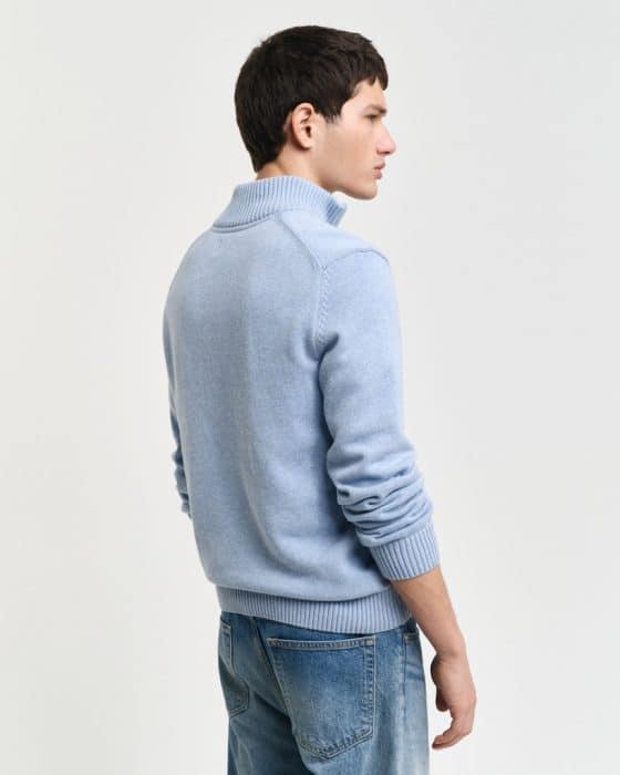 A person wearing a light blue, long-sleeve sweater and blue jeans stands against a plain white background. The person is facing sideways, showing the sweaters ribbed neckline and cuffs.