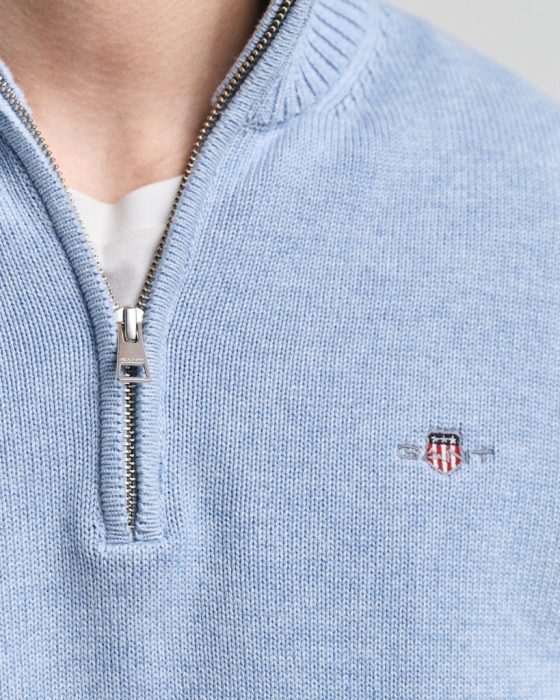 Close-up of a person wearing a light blue sweater with a quarter zipper. A small embroidered crest with red and white stripes and a subtle letter design is visible on the chest. The zipper is partially opened, revealing a white shirt underneath.