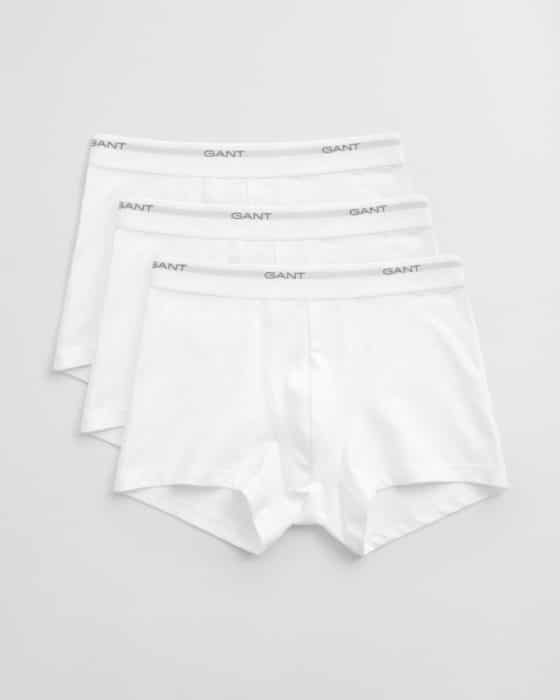 Three pairs of white boxer briefs are laid out in a slightly overlapping arrangement. Each pair has an elastic waistband with the brand name GANT printed in gray. The background is a plain, light gray surface.