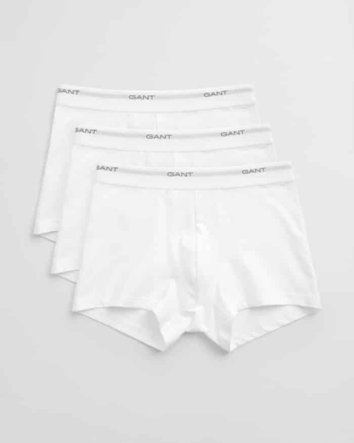 Three pairs of white boxer briefs are laid out in a slightly overlapping arrangement. Each pair has an elastic waistband with the brand name GANT printed in gray. The background is a plain, light gray surface.