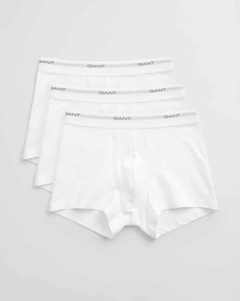 Three pairs of white boxer briefs are laid out in a slightly overlapping arrangement. Each pair has an elastic waistband with the brand name GANT printed in gray. The background is a plain, light gray surface.