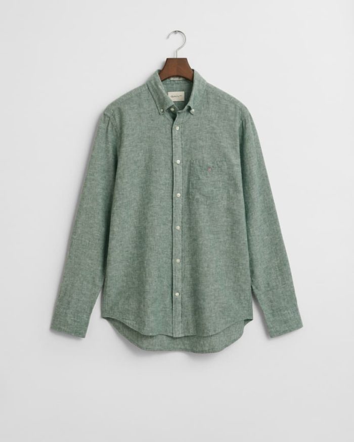 A long-sleeved, button-up green shirt with a textured fabric hangs on a wooden hanger against a white background. The shirt features a left chest pocket and a collared neckline.