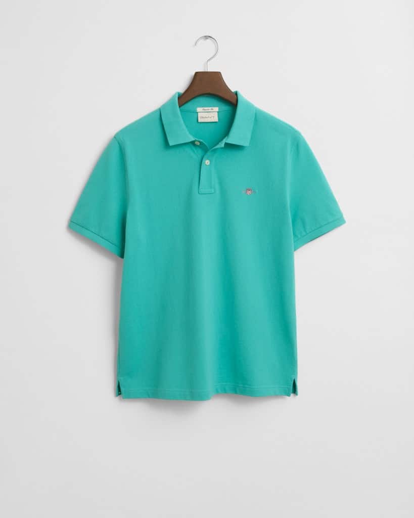 A teal polo shirt hangs on a hanger against a plain white background. The shirt has a collar, short sleeves, and a small embroidered logo on the upper left side.