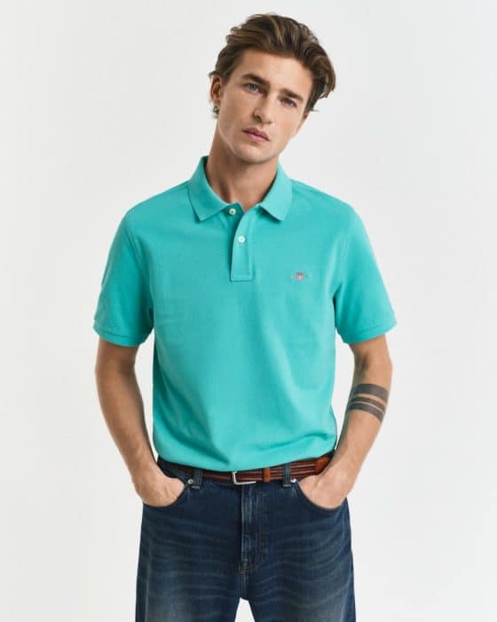 A person wearing a turquoise polo shirt and dark jeans stands with hands in pockets, looking ahead. They have short brown hair and a tattoo on one forearm.