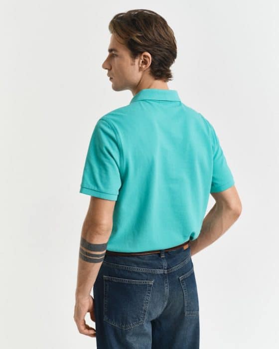 A person wearing a teal polo shirt and dark jeans is standing and facing left. They have short brown hair and an arm tattoo. The background is a plain, light-colored wall.