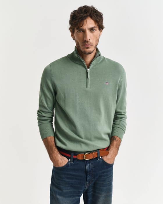 A person with short curly hair stands against a plain background, wearing a light green quarter-zip sweater and blue jeans. They have their hands in their pockets and are looking directly at the camera.