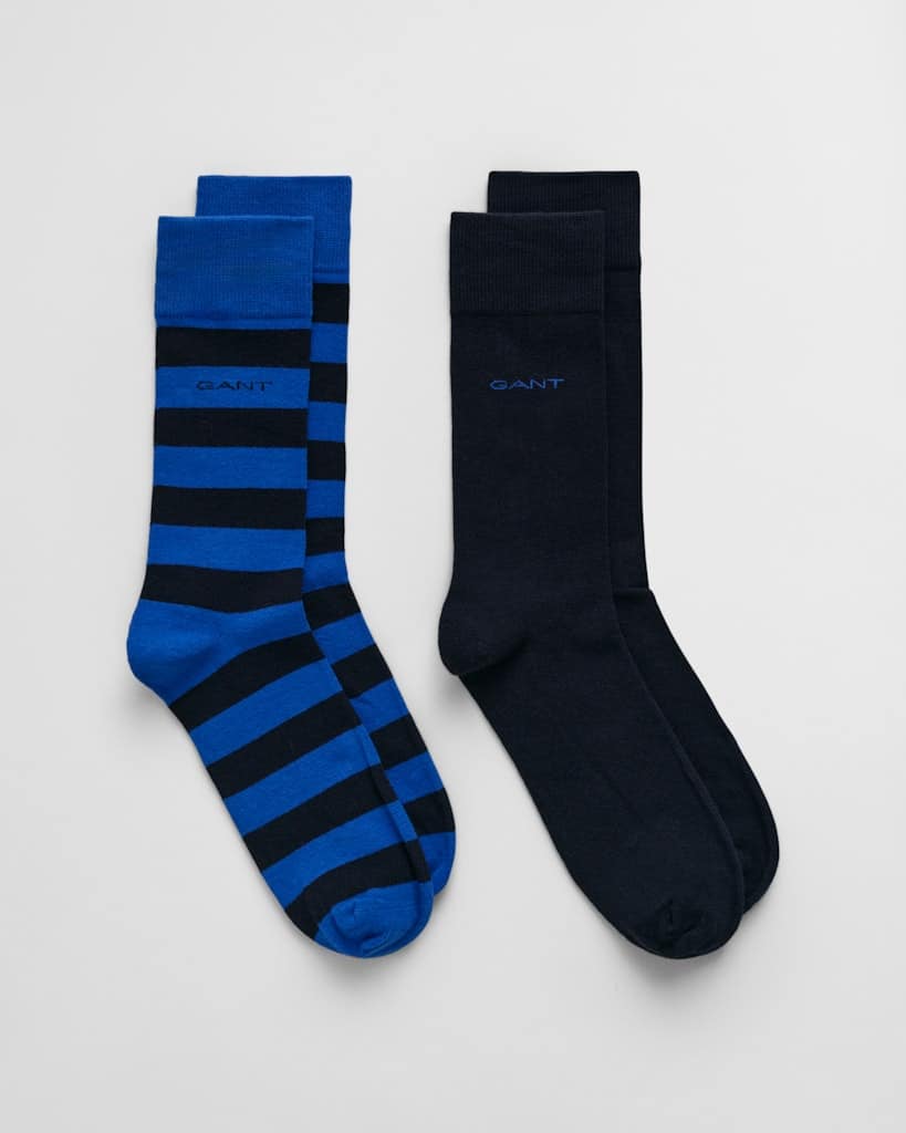 Two pairs of crew socks are displayed. One pair features blue and black horizontal stripes, while the other is solid black. Both pairs have the brand name GANT embroidered near the top. The socks are laid flat on a light background.