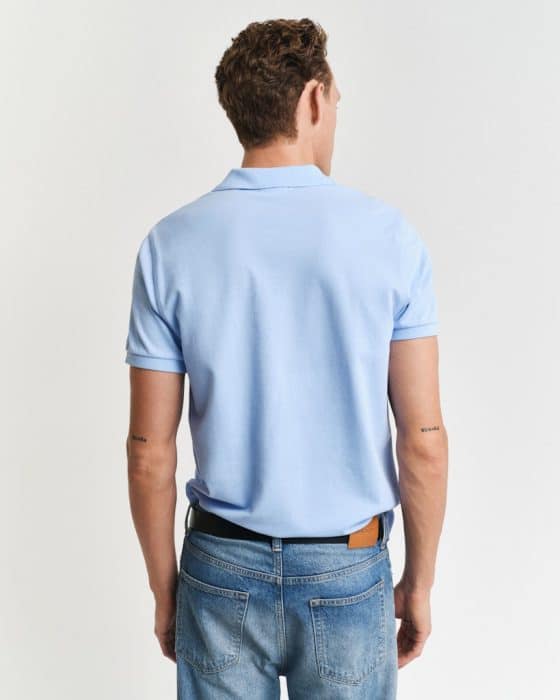 A man with short curly hair is standing with his back to the camera. He is wearing a light blue polo shirt and blue jeans, with a brown belt. Two small tattoos are visible on his forearms. The background is plain white.