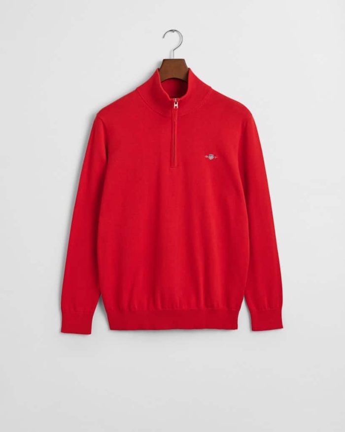 A red pullover with a half-zip collar is hanging on a wooden hanger. It features a small embroidered logo on the left chest. The background is a plain white wall.