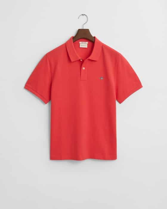 A red polo shirt with short sleeves hangs on a wooden hanger against a plain white background. It features a small embroidered logo on the left chest.