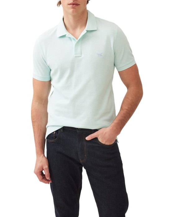 A person wearing a light green short-sleeve polo shirt with a small logo on the chest, paired with dark jeans. The individual is standing with one hand in their pocket. The background is plain white.