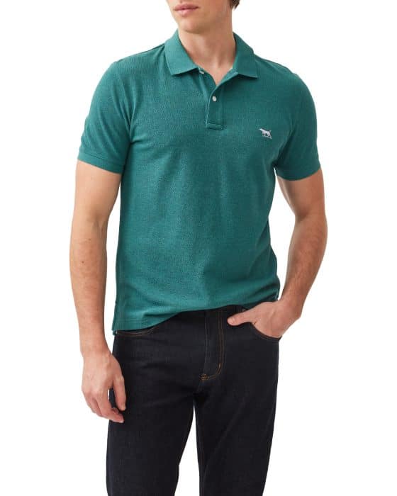 A person wearing a teal polo shirt with a small emblem on the chest and dark jeans. The person has one hand in their pocket and is standing against a white background.