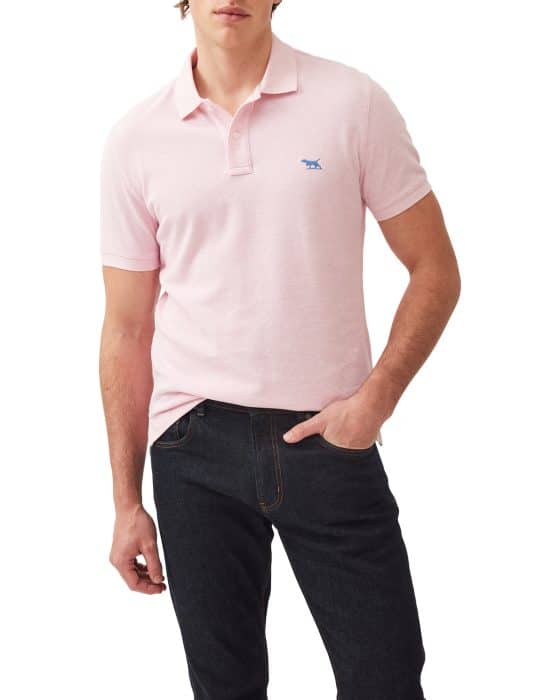 A person wearing a light pink polo shirt with a small blue embroidered logo and dark jeans. The shirt has short sleeves and a collar. The person stands with one hand in their pocket, posing against a plain white background.