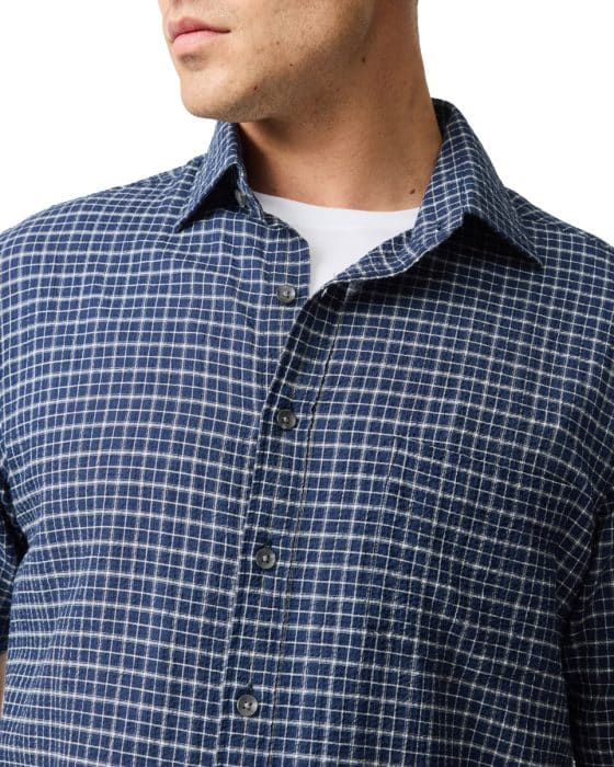 Person wearing a blue checkered button-up shirt with a white undershirt, facing slightly to the left. The shirt has a small chest pocket and a classic collar. The focus is on the upper body and clothing details.