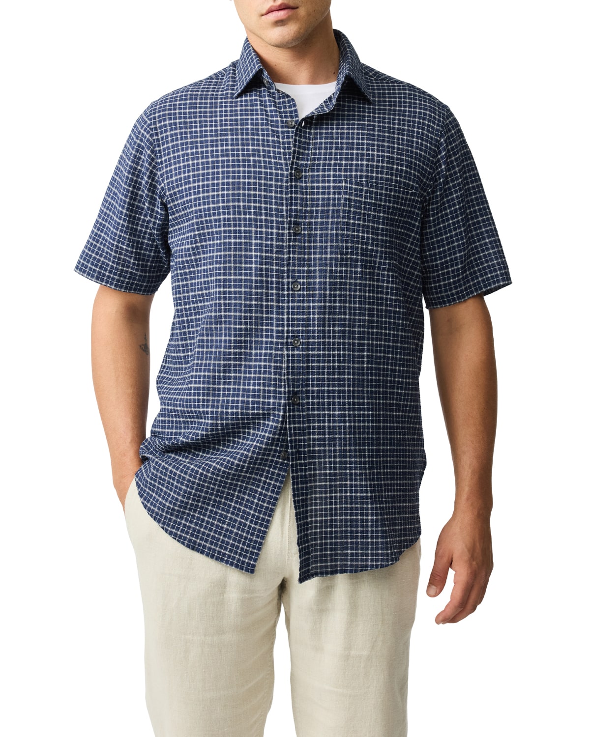 A person wearing a blue checkered short-sleeve button-up shirt over a white t-shirt and light beige pants. The persons hand is in their pocket. The outfit has a casual, relaxed style.