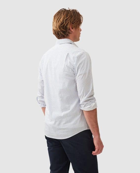 A person with light brown hair is seen from the back, wearing a white, long-sleeved, patterned shirt and dark pants against a plain gray background. Their arms are at their sides.