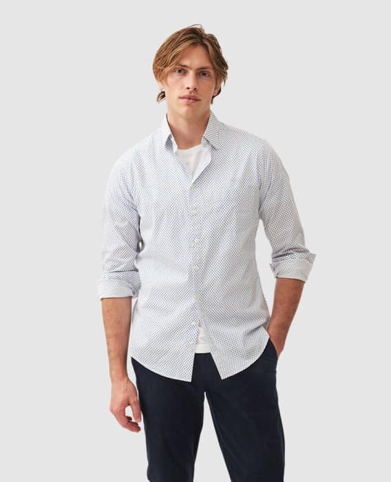 A person with medium-length hair wears a white patterned button-up shirt over a white t-shirt and dark pants. The sleeves are rolled up. The background is plain gray.