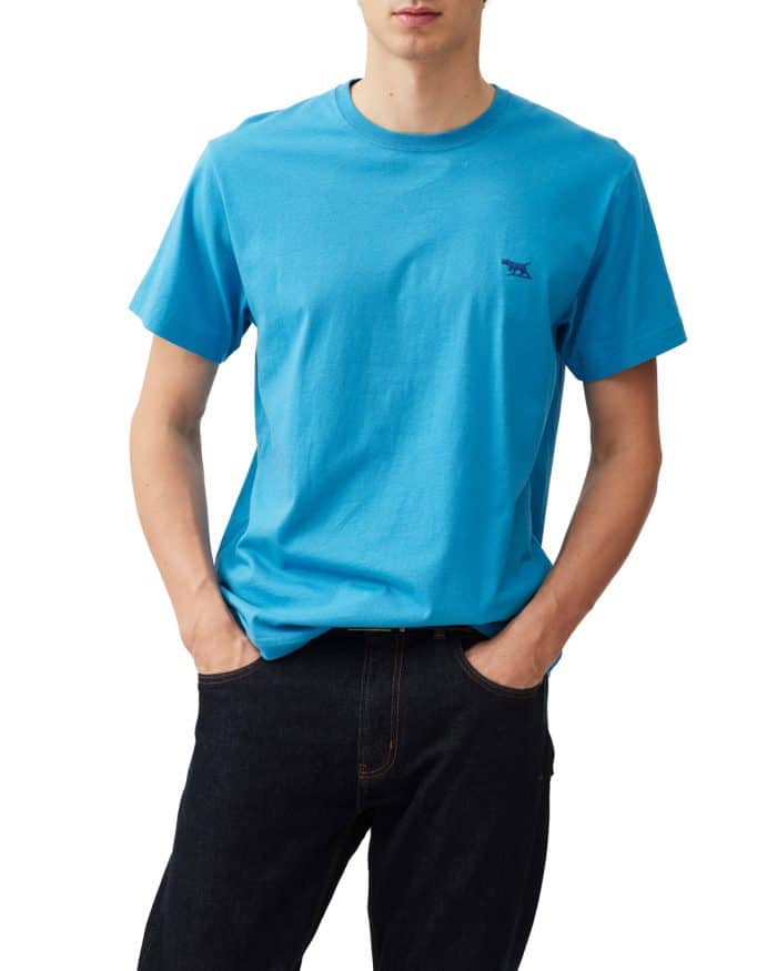 A person wearing a bright blue T-shirt with a small logo on the chest stands with hands in the pockets of black jeans. The background is plain white.