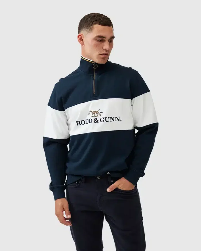 A man is wearing a navy blue and white Rodd & Gunn pullover with a quarter zip. The shirt features the brands logo with a dog design. He has short hair and is posing against a plain light gray background.