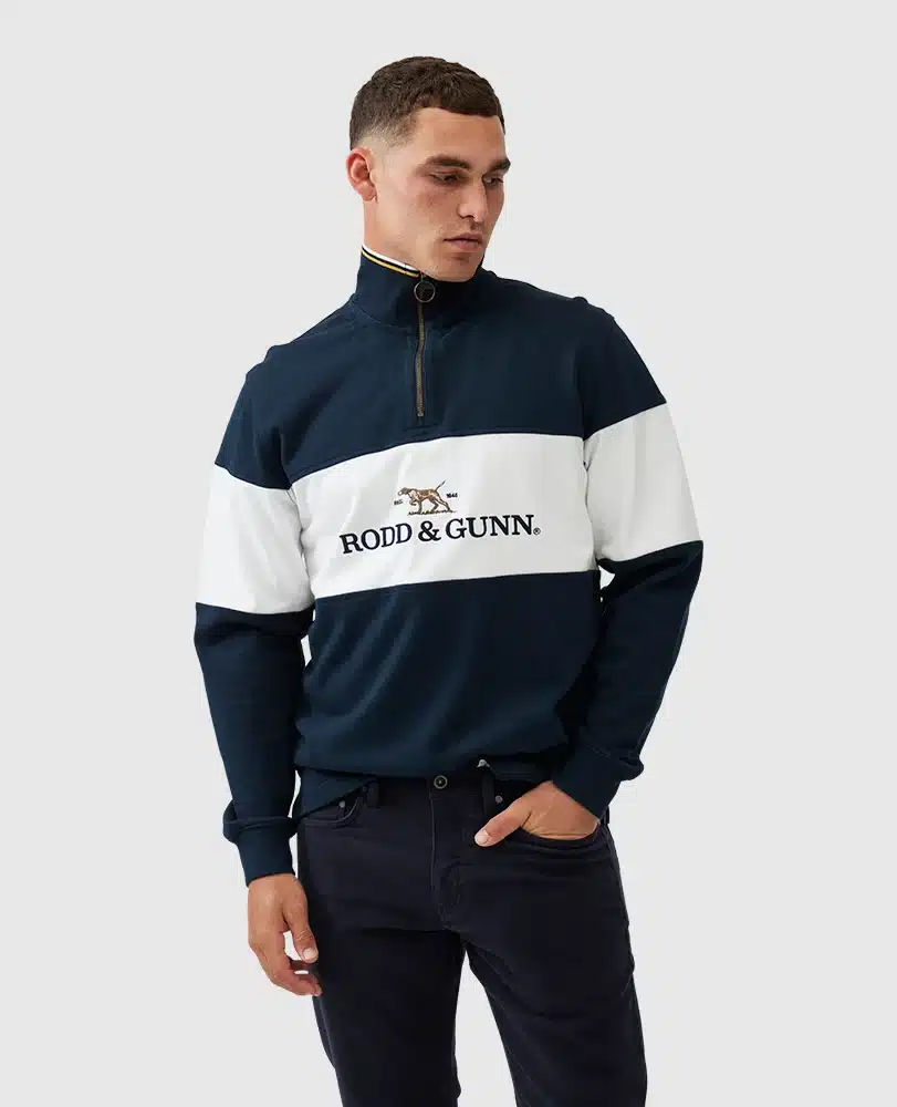 A man is wearing a navy blue and white Rodd & Gunn pullover with a quarter zip. The shirt features the brands logo with a dog design. He has short hair and is posing against a plain light gray background.