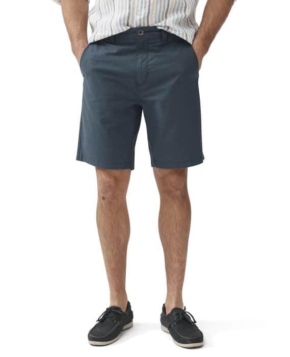 A person wearing a striped shirt, teal shorts, and dark boat shoes stands with hands in pockets. The background is plain white.