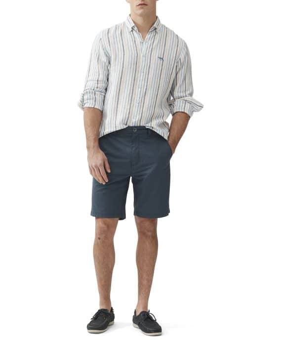 A person wearing a light blue and white striped shirt with rolled-up sleeves, dark blue shorts, and dark loafers stands against a plain white background. Their hands are relaxed, one in the pocket.