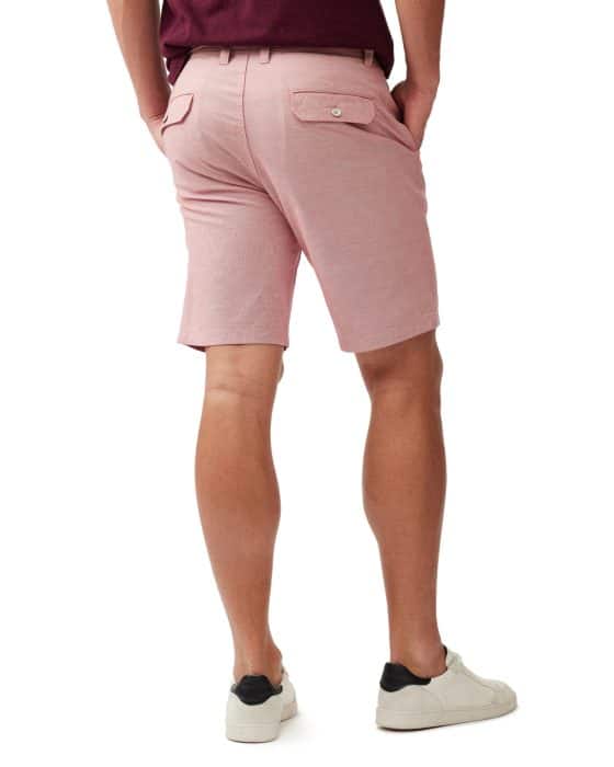 A person wearing a maroon shirt, pink shorts with back pockets, and white sneakers is standing with hands in pockets, facing away from the camera. The background is plain white.