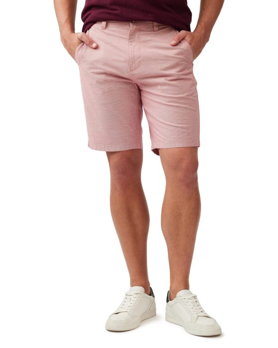 Man wearing pink shorts, with hands in pockets, wears a burgundy shirt tucked in. He is also wearing white sneakers with black accents. The background is plain white.