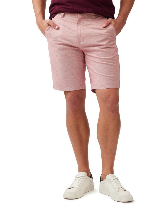 Man wearing pink shorts, with hands in pockets, wears a burgundy shirt tucked in. He is also wearing white sneakers with black accents. The background is plain white.