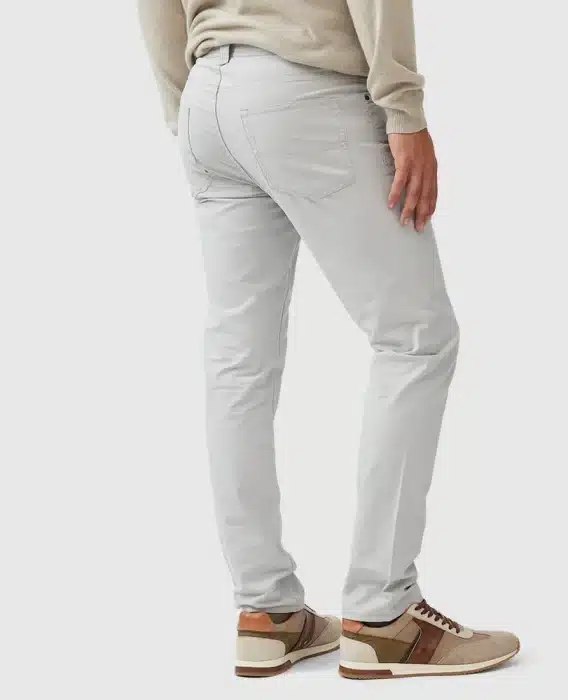 A person wearing light gray slim-fit jeans and beige-brown sneakers is standing with one leg slightly forward. They are also wearing a light-colored sweater, with the focus on the lower body. The background is plain white.