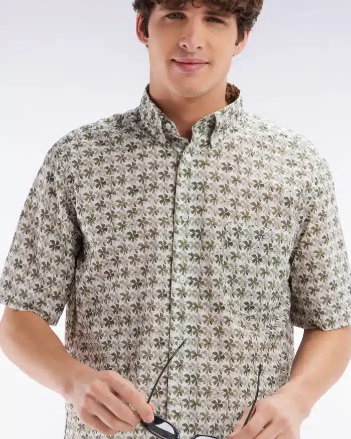 Man in a patterned, short-sleeve button-up shirt holds glasses. He has curly brown hair and smiles slightly. The shirt features a green and brown floral design. The background is a simple gradient from white to light gray.