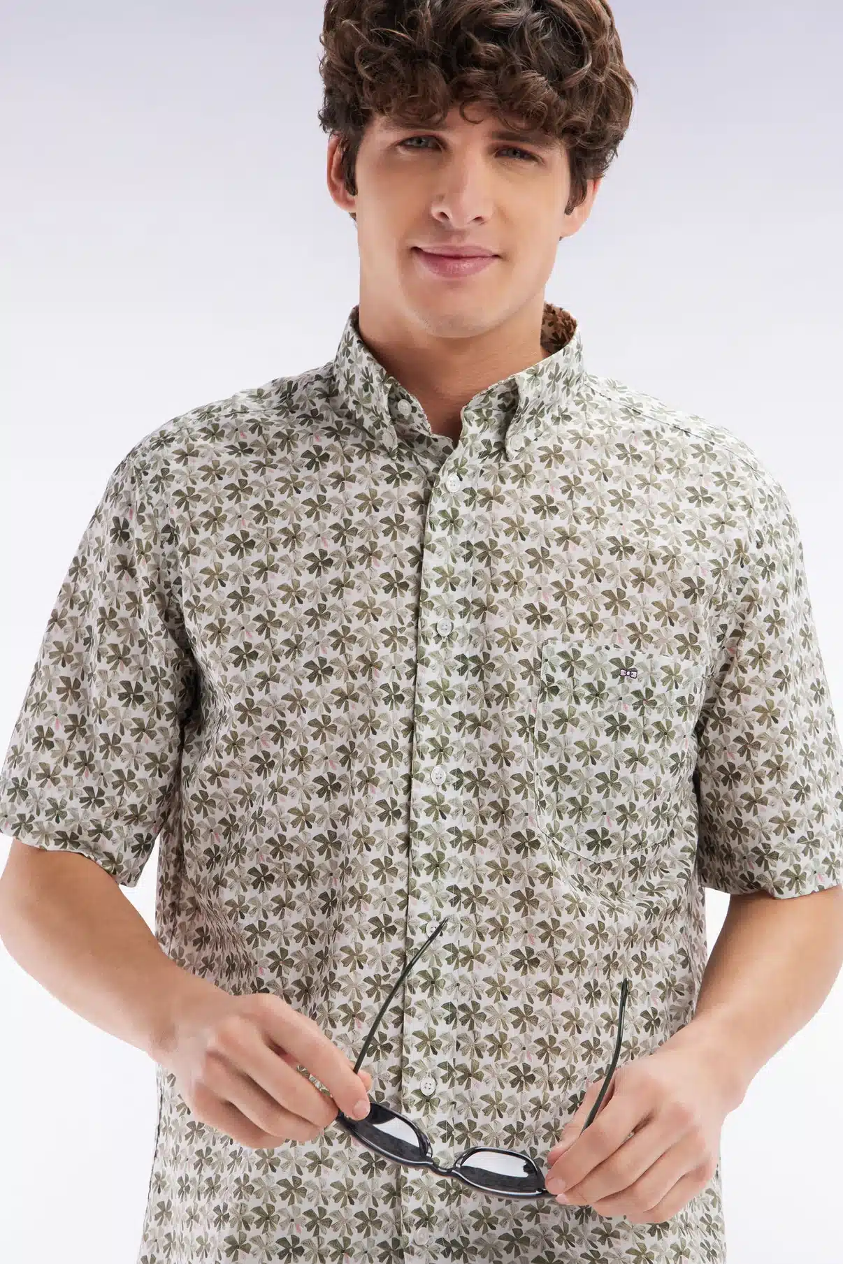 Man in a patterned, short-sleeve button-up shirt holds glasses. He has curly brown hair and smiles slightly. The shirt features a green and brown floral design. The background is a simple gradient from white to light gray.