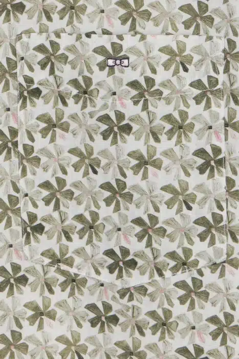 Close-up of a fabric pattern with green abstract floral shapes on a white background. There is a pocket in the center featuring a small label with text. The design has accents of pink and beige within the green shapes.