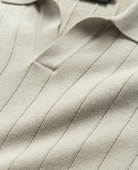 Close-up of a beige knit polo shirt featuring a subtle vertical ribbed pattern and a partially open collar. The fabric appears soft and textured, highlighting its fine knit quality.