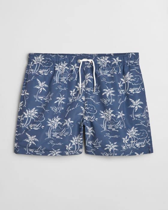 Blue swim trunks with a white tropical print featuring palm trees, surfers, and script writing. The shorts have an elastic waistband and a white drawstring.