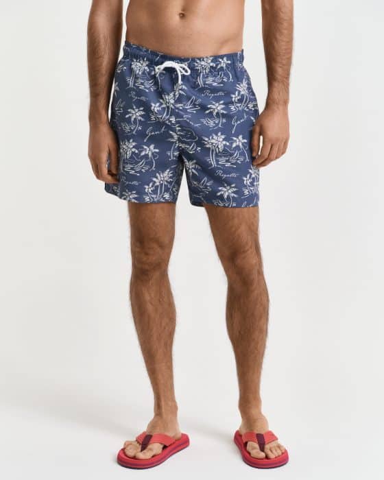 A person wearing navy blue swim shorts with a white floral pattern and script design, paired with bright red flip-flops. The image is cropped at the waist and below the shoulders, showing the legs and feet against a plain white background.