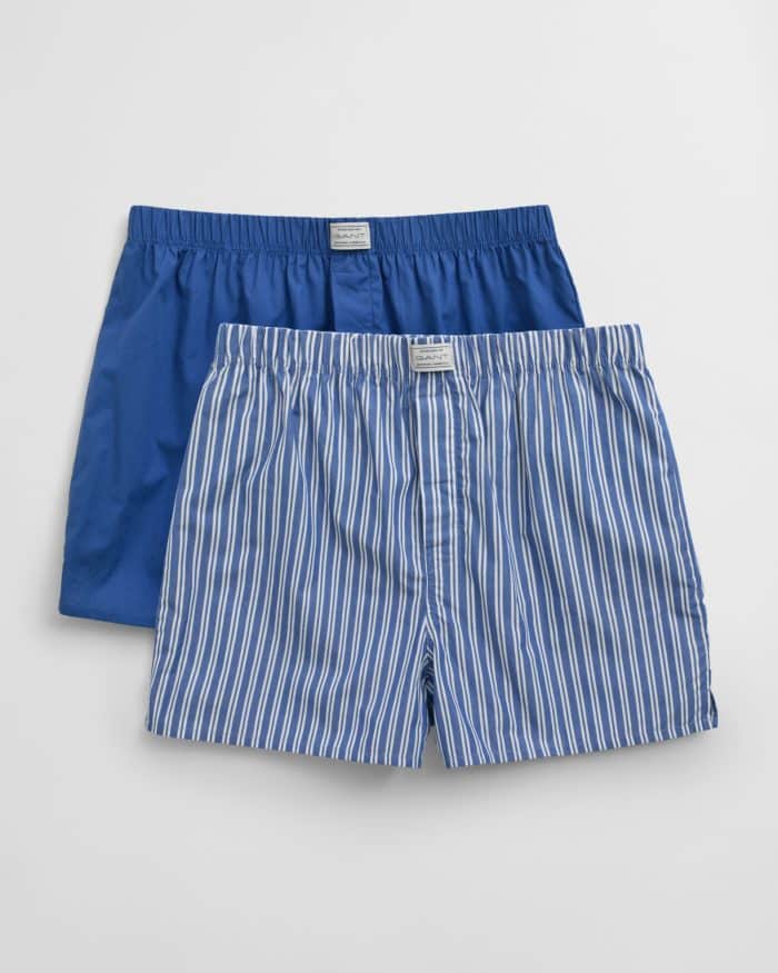 Two pairs of mens boxer shorts on a white background. One pair is solid blue and the other pair has a blue and white vertical stripe pattern. Both have elastic waistbands and a button fly.