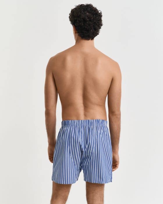 A person with curly hair is standing with their back to the camera, wearing blue and white striped boxer shorts. They are shirtless, and the background is plain white.