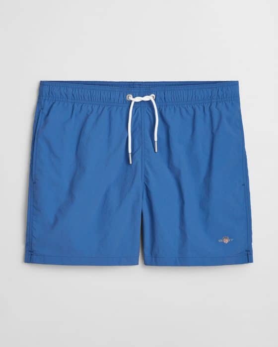 Blue swim shorts with an elastic waistband and a white drawstring. A small embroidered logo is visible on one leg. The fabric appears lightweight and suitable for swimming. Shorts are laid flat on a plain background.